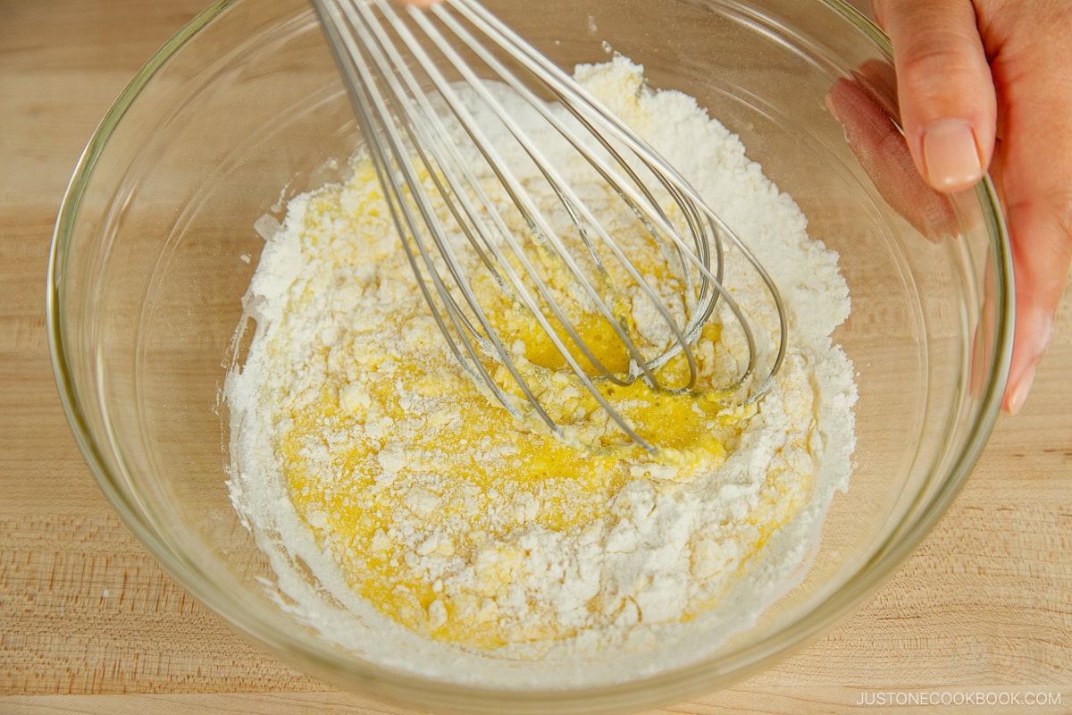 A hand whisk in a glass mixing bowl containing batter and flour for fluffy Japanese soufflé pancakes.